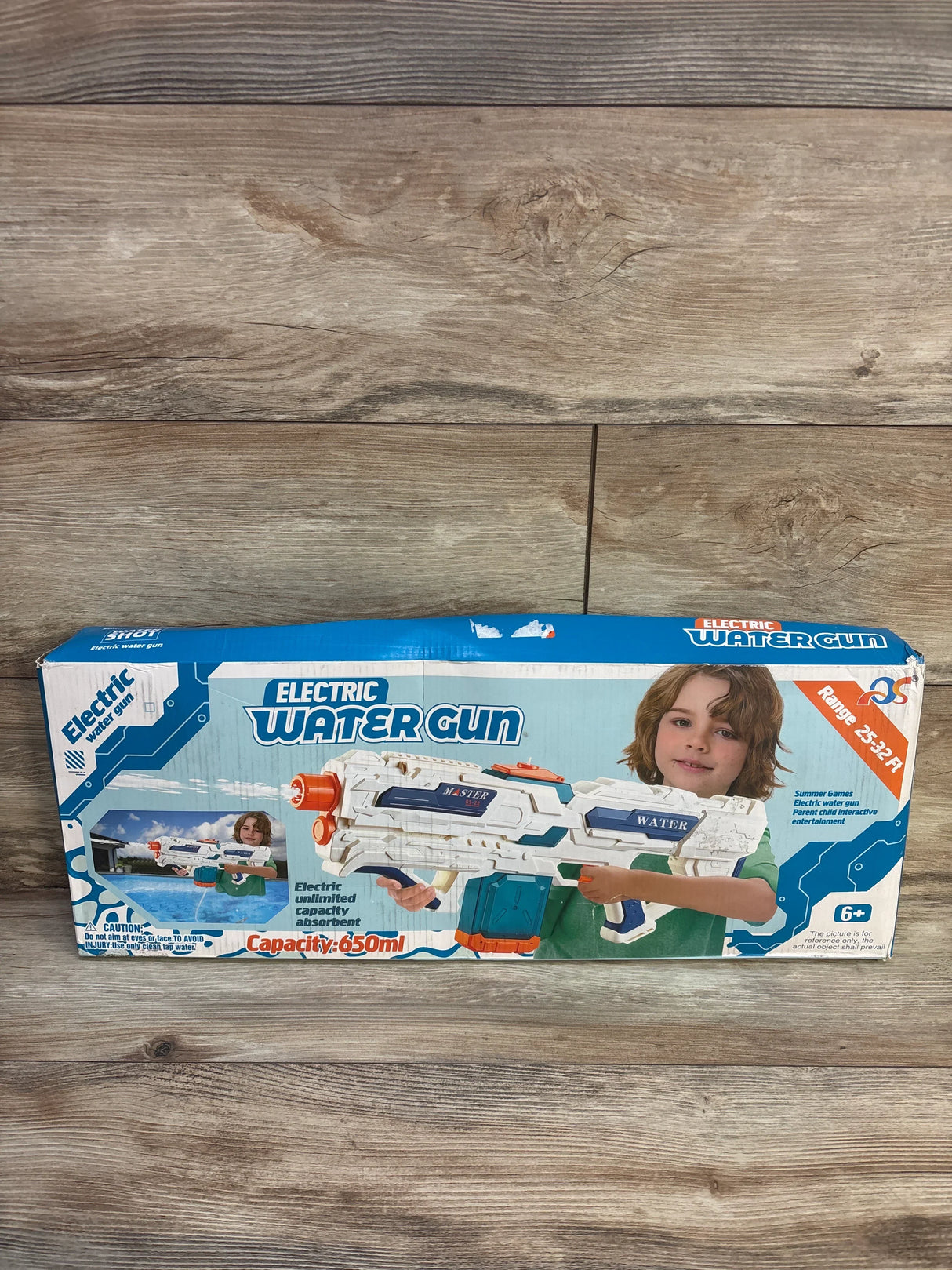Front view of electric water cannon box showing white-blue water gun, 650ml capacity and 25-32 ft range on wood background
