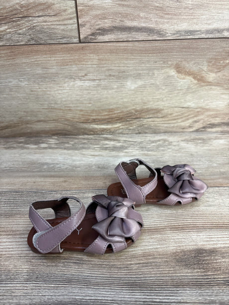 Front three-quarter view of EYY mauve baby sandals showing large bow detail and Velcro ankle straps on wood background