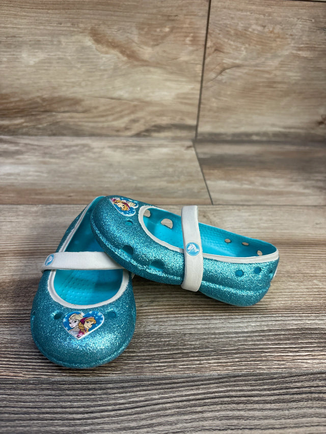 Front and side view of kids' Crocs Frozen Mary Jane flats in blue glitter showcasing white Mary Jane strap and Frozen character heart charm on wood floor background