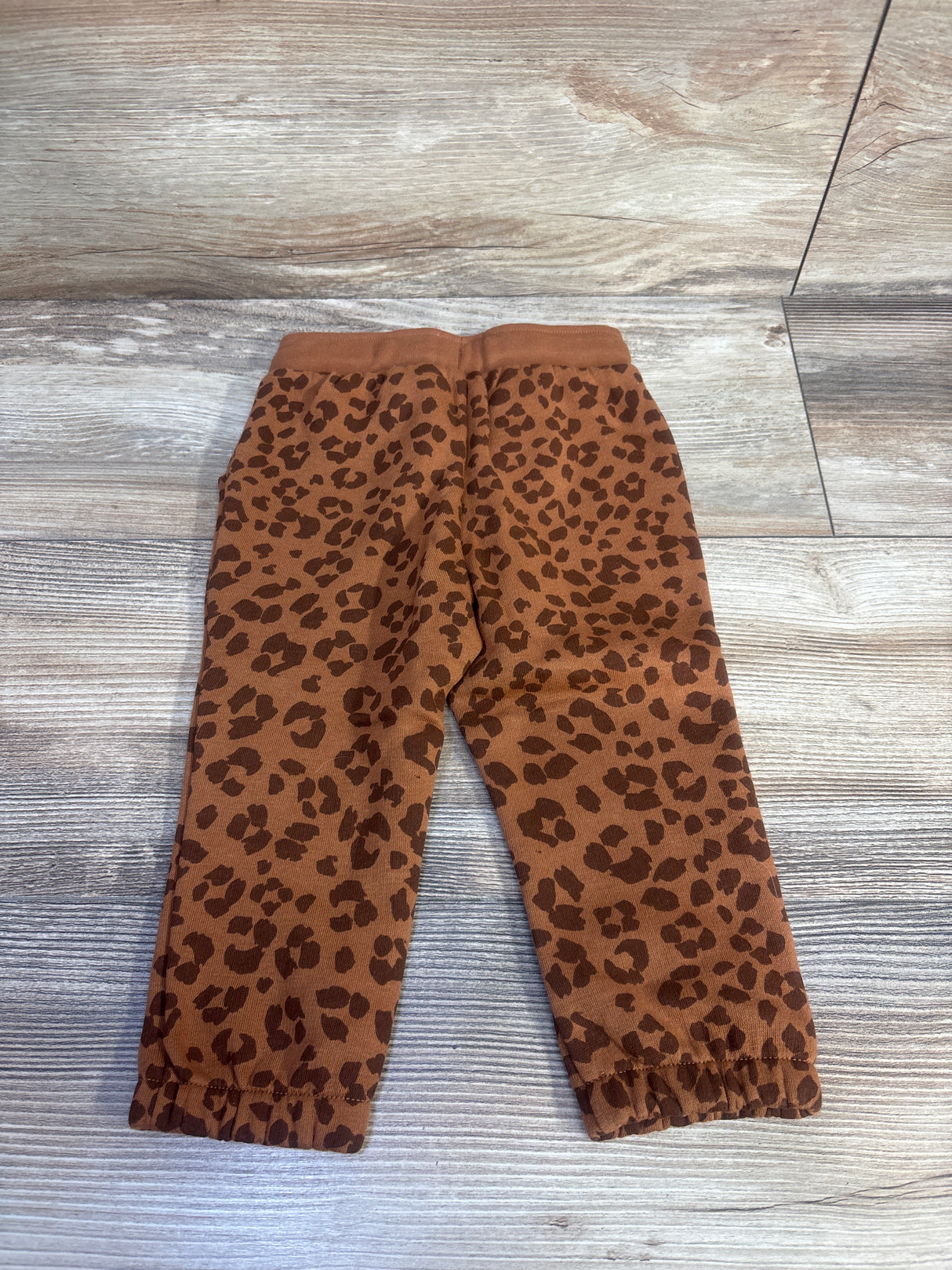 Flat lay of brown leopard print Okie Dokie jogger pants showing elastic waistband and cuffed ankles on light wood background