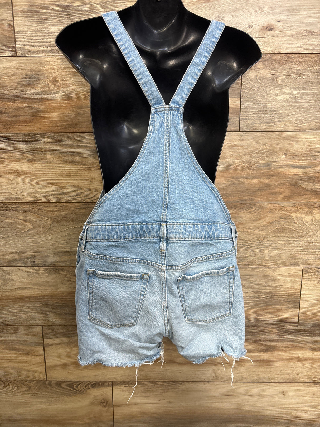 Back view of Isabel maternity denim shortalls in light blue showing frayed hem, two back pockets and crossed straps on a black mannequin against wood background