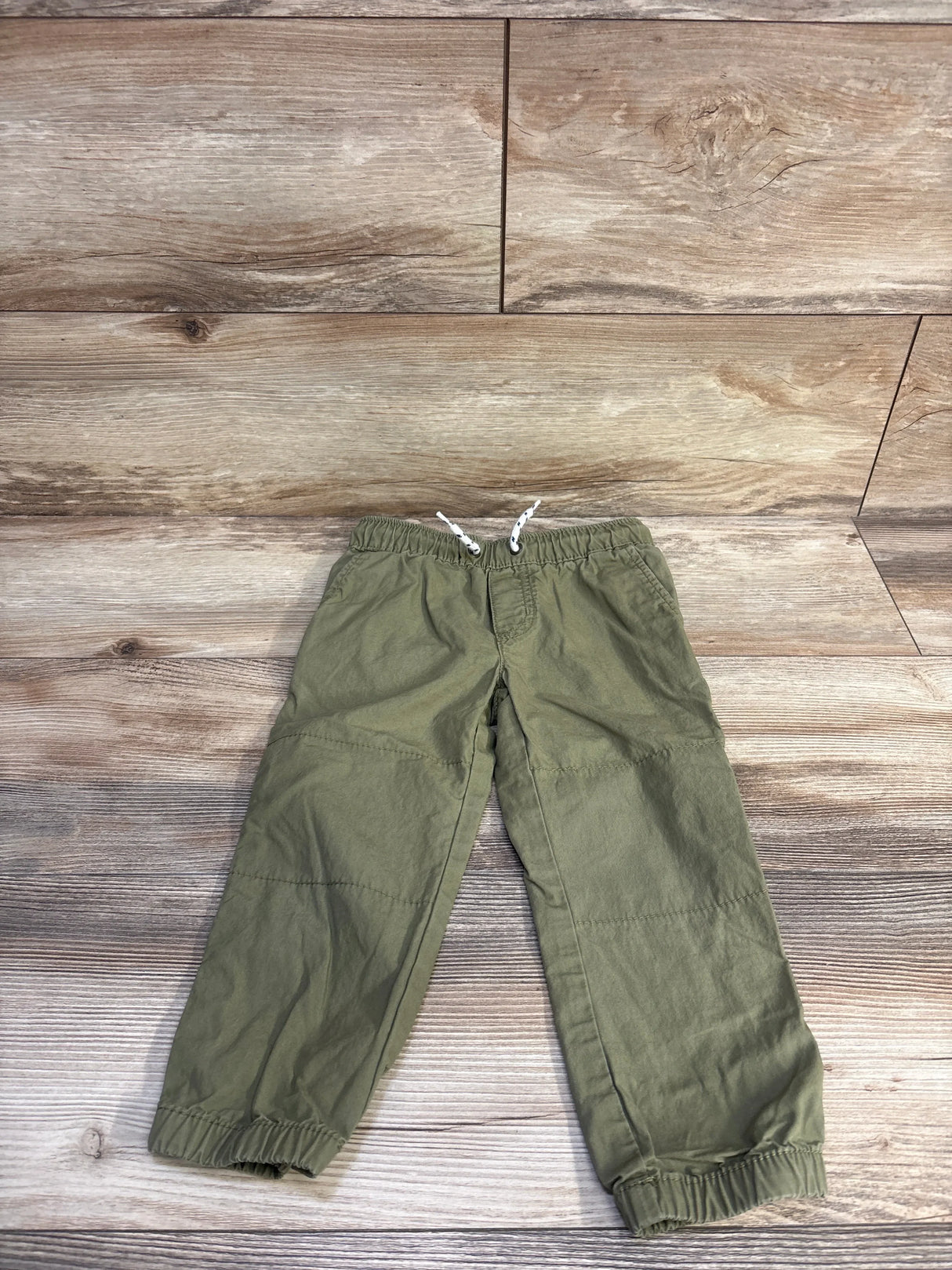 Flat lay of green toddler drawstring pants showcasing elastic waistband with white drawstring and elastic cuffs on wood background