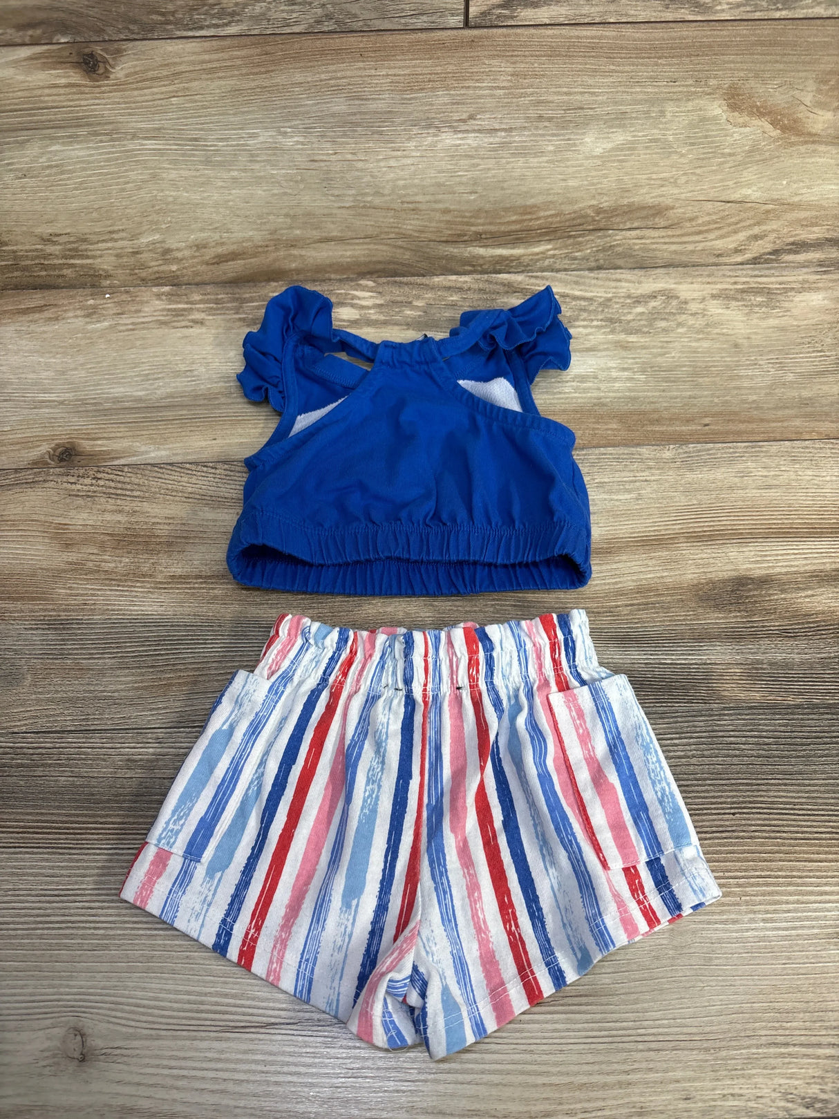 Flat lay of girls' 2-piece Fourth of July set showcasing ruffled blue top and elastic-waist red white and blue striped shorts on wood background