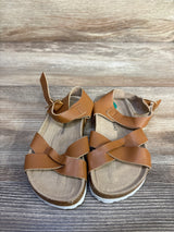 Top-down view of Lucky Brand Kiara toddler sandals, brown; crossed straps, ankle Velcro, contoured footbed on wood background