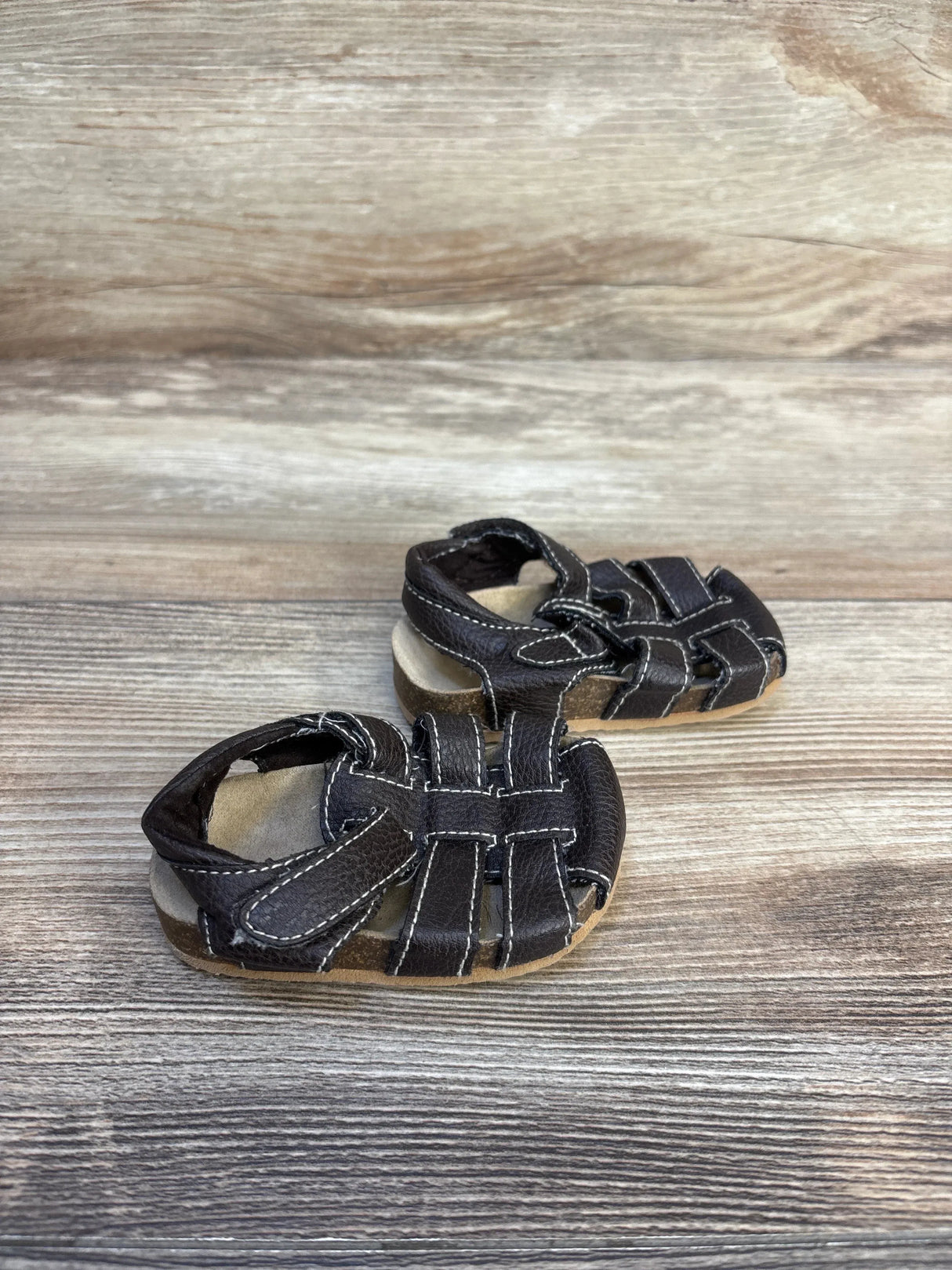Side view of Children's Place leather fisherman sandals in dark brown with white stitching and hook and loop strap on wood plank floor
