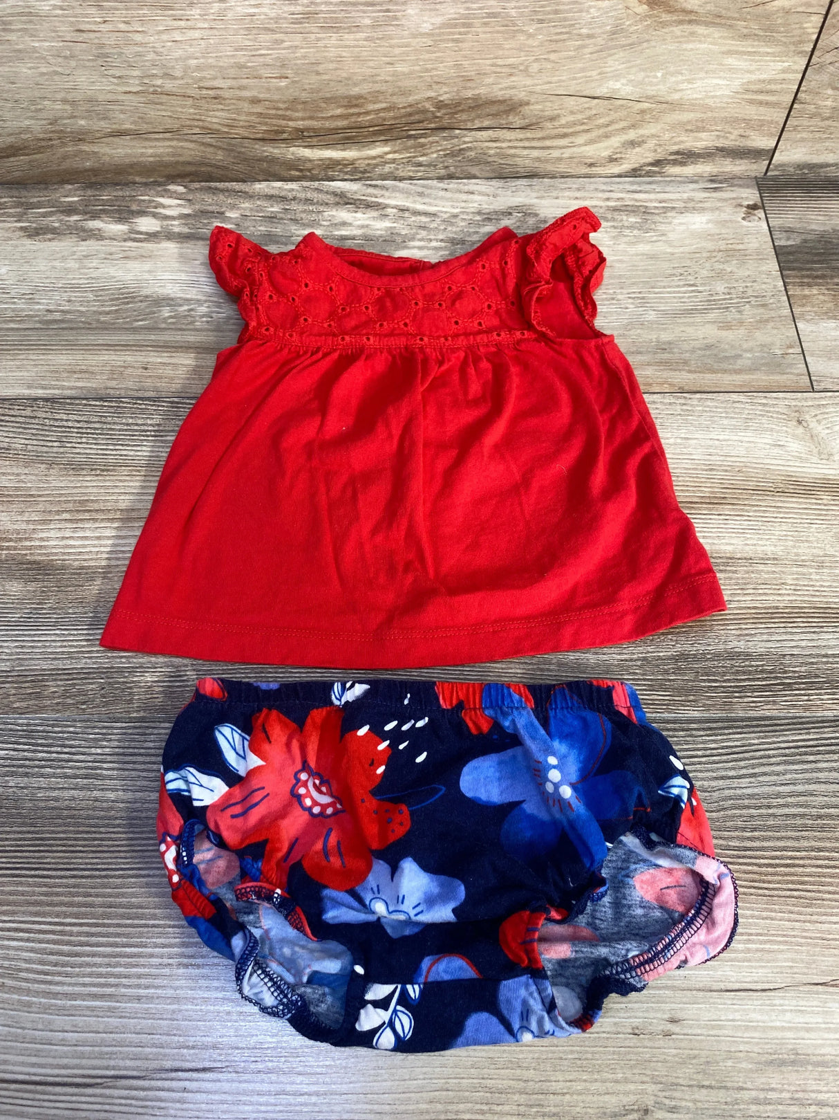 Flat lay of Carter's 2-piece red eyelet top and navy floral bloomers showing eyelet yoke and flutter sleeves with elastic waist on light wood background