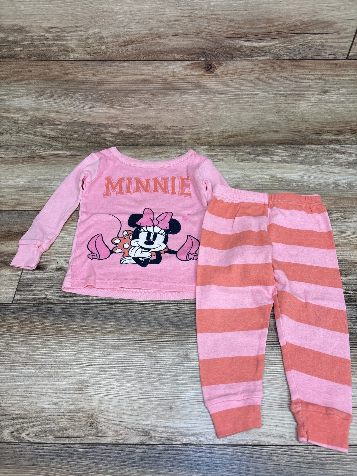 Front view of Disney Junior Minnie Mouse two-piece pink pajama set with Minnie graphic and striped pants on wood background