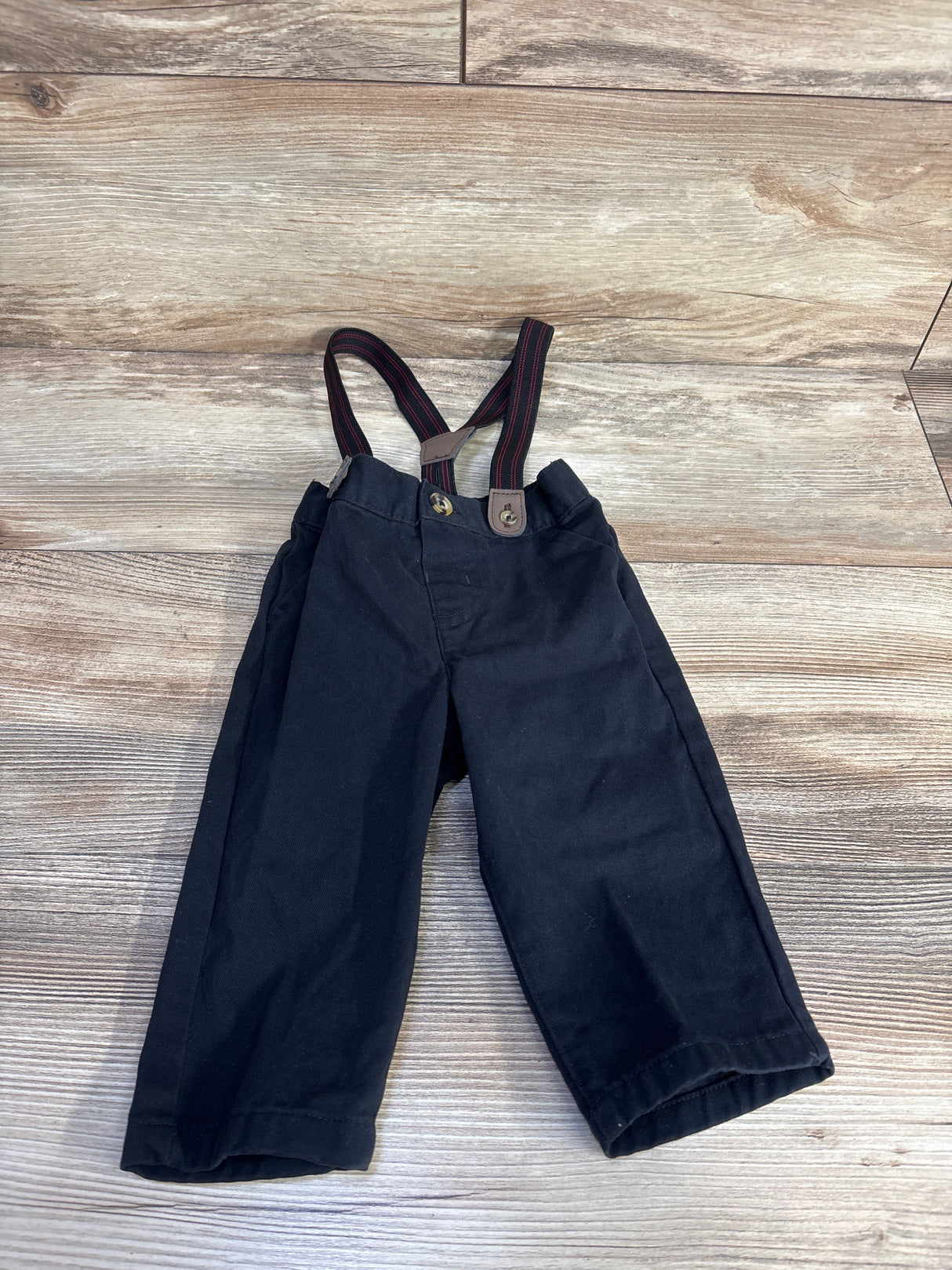 Top-down view of black baby suspender pants showing red-striped elastic suspenders and front button tabs on a light wood floor