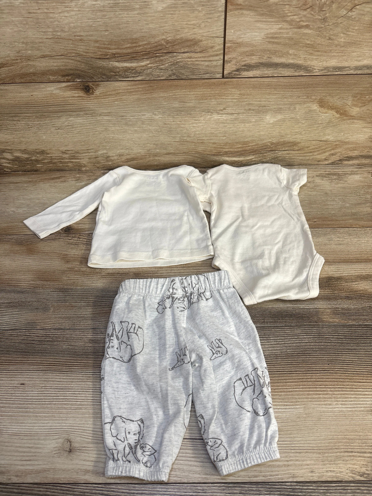 Flat lay of Carter's 3-piece ivory set: long sleeve top, short sleeve bodysuit, and gray elephant-print pants on wood floor