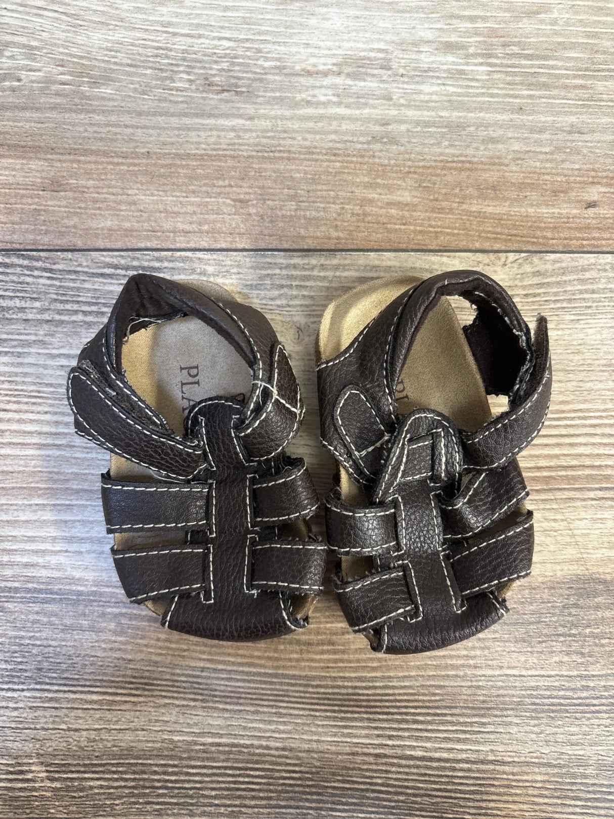 Brown leather toddler sandals with white stitching on wood floor