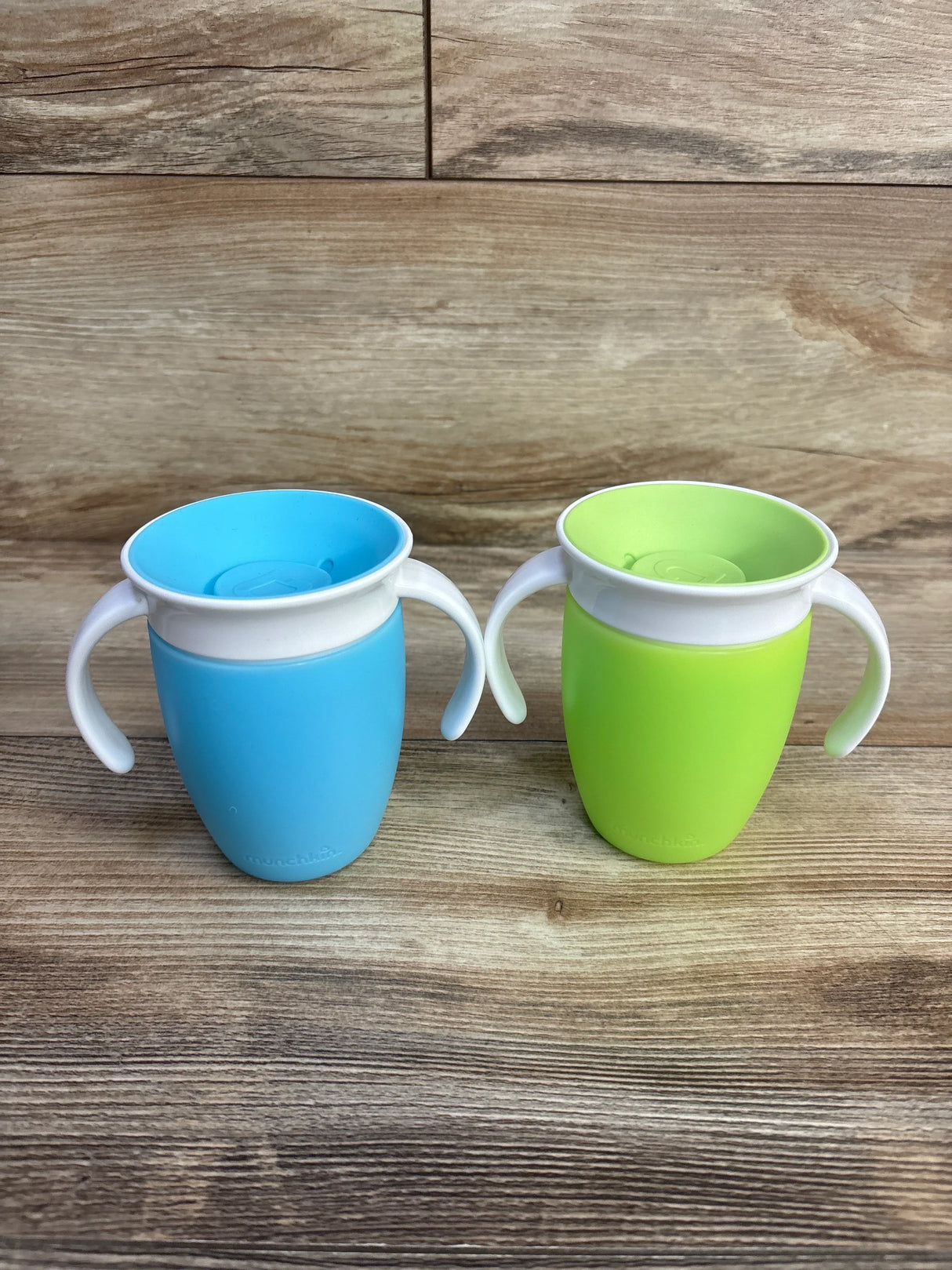Front view of two Munchkin Miracle 360 trainer cups, blue and green, showing handles and 360-degree spill-proof rims on wood background