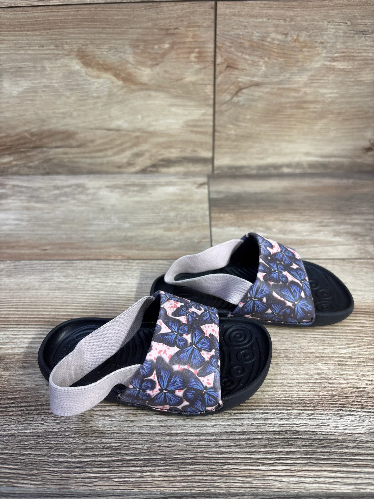 Angled view of Nike toddler Kawa slides with blue butterfly strap, elastic heel strap, and black footbed on wood background