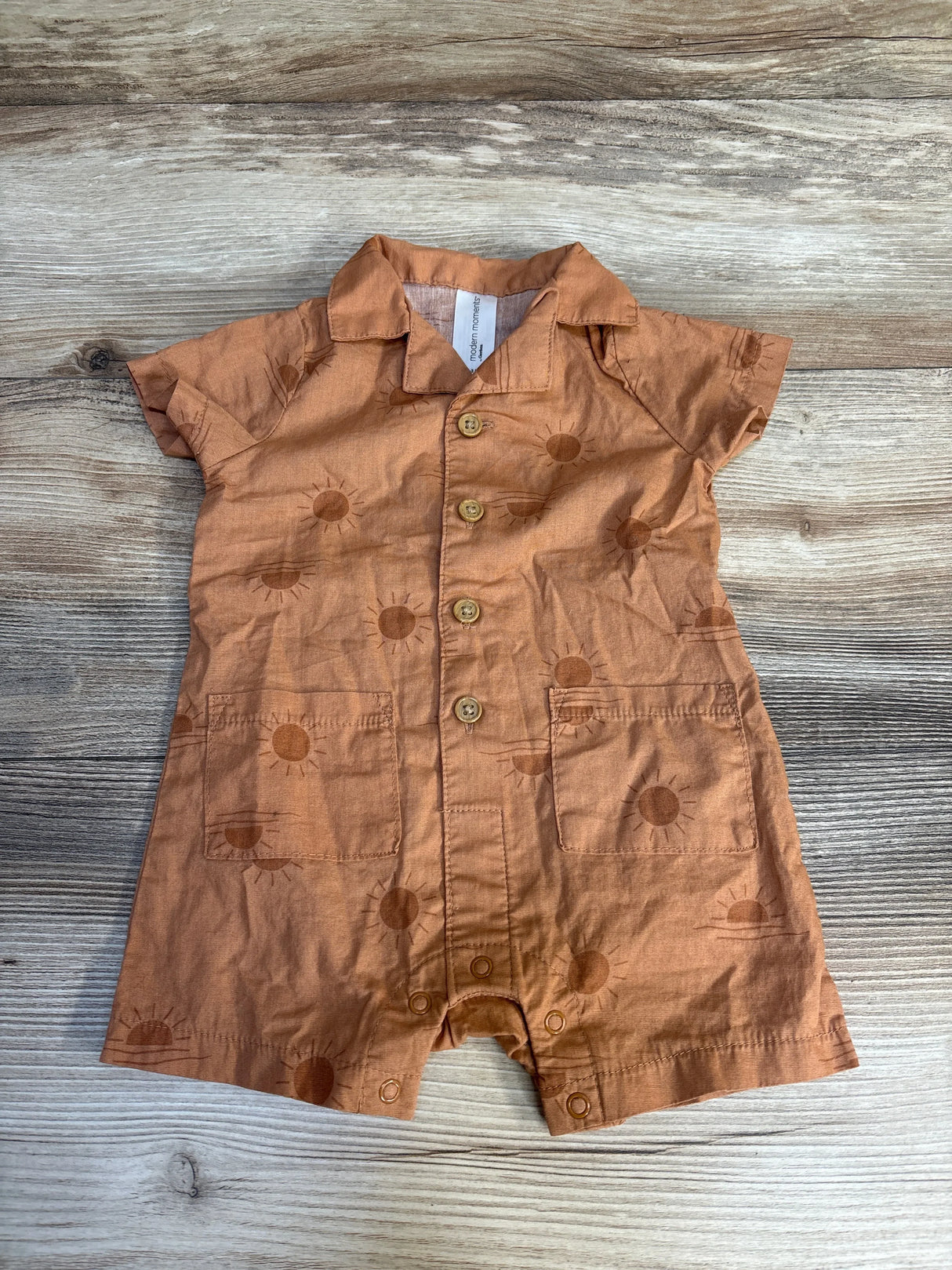 Rust-colored baby romper with sun pattern, short sleeves, and front buttons on wood background.