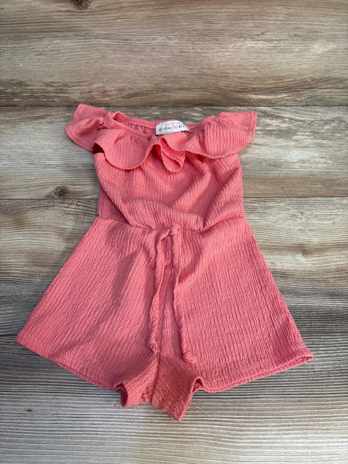 Pink ruffle off-shoulder toddler romper with tie waist, laid flat on wooden surface