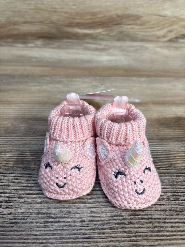Pink knit baby booties with unicorn horns, ears, and smiling faces on wood floor