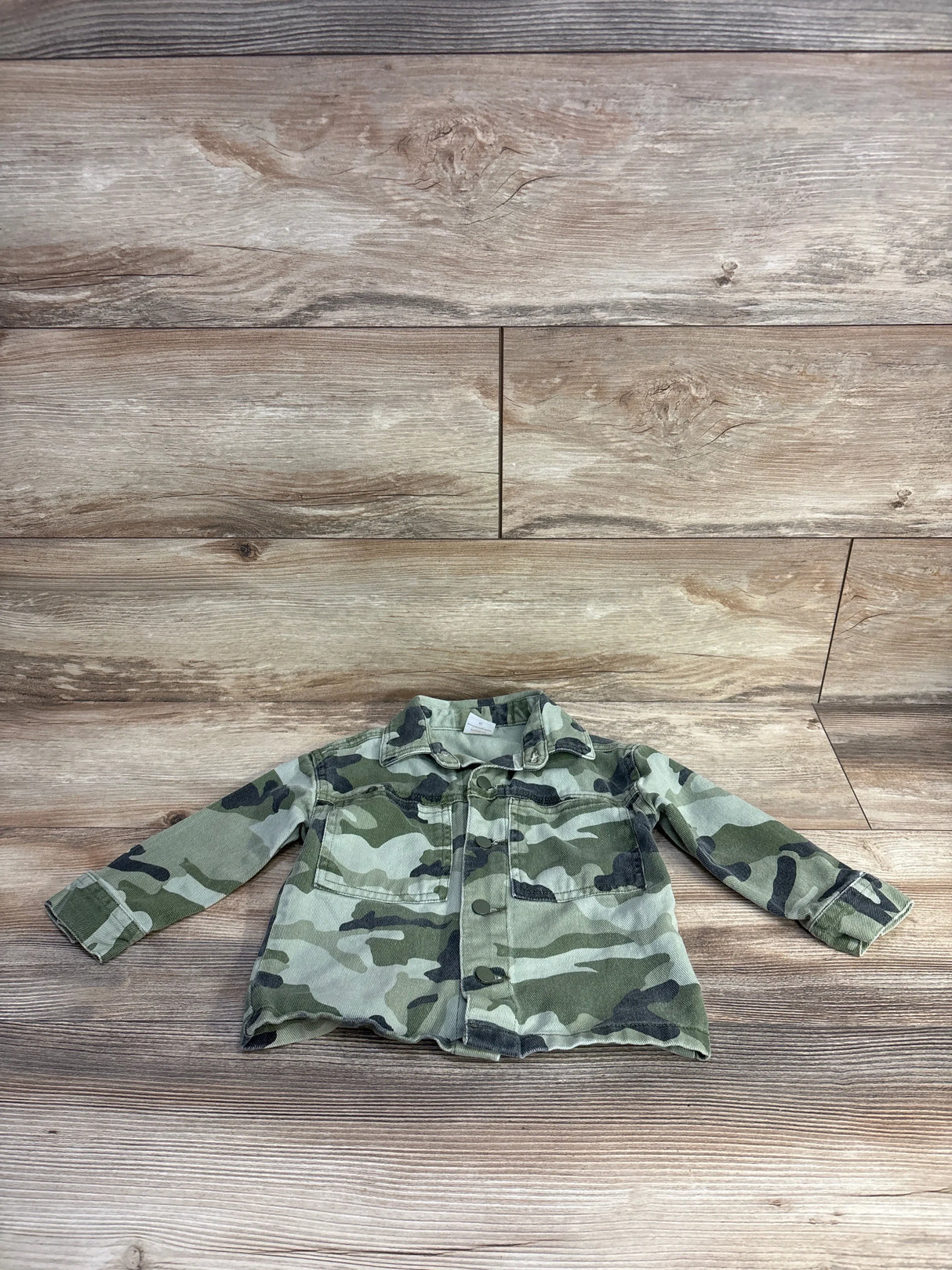 Front view of toddler camo denim jacket size 2T showcasing button-front closure and chest pockets with wood plank background