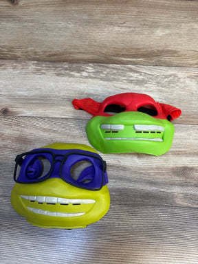 Two Teenage Mutant Ninja Turtles kids masks, one with a red bandana and one with purple glasses, on wood background.