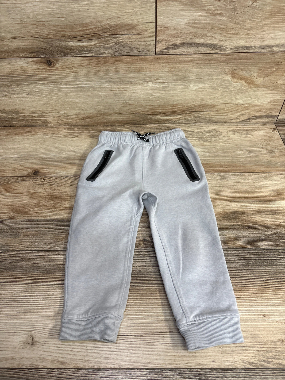 Light gray toddler sweatpants with black zippered pockets and elastic cuffs on wooden floor