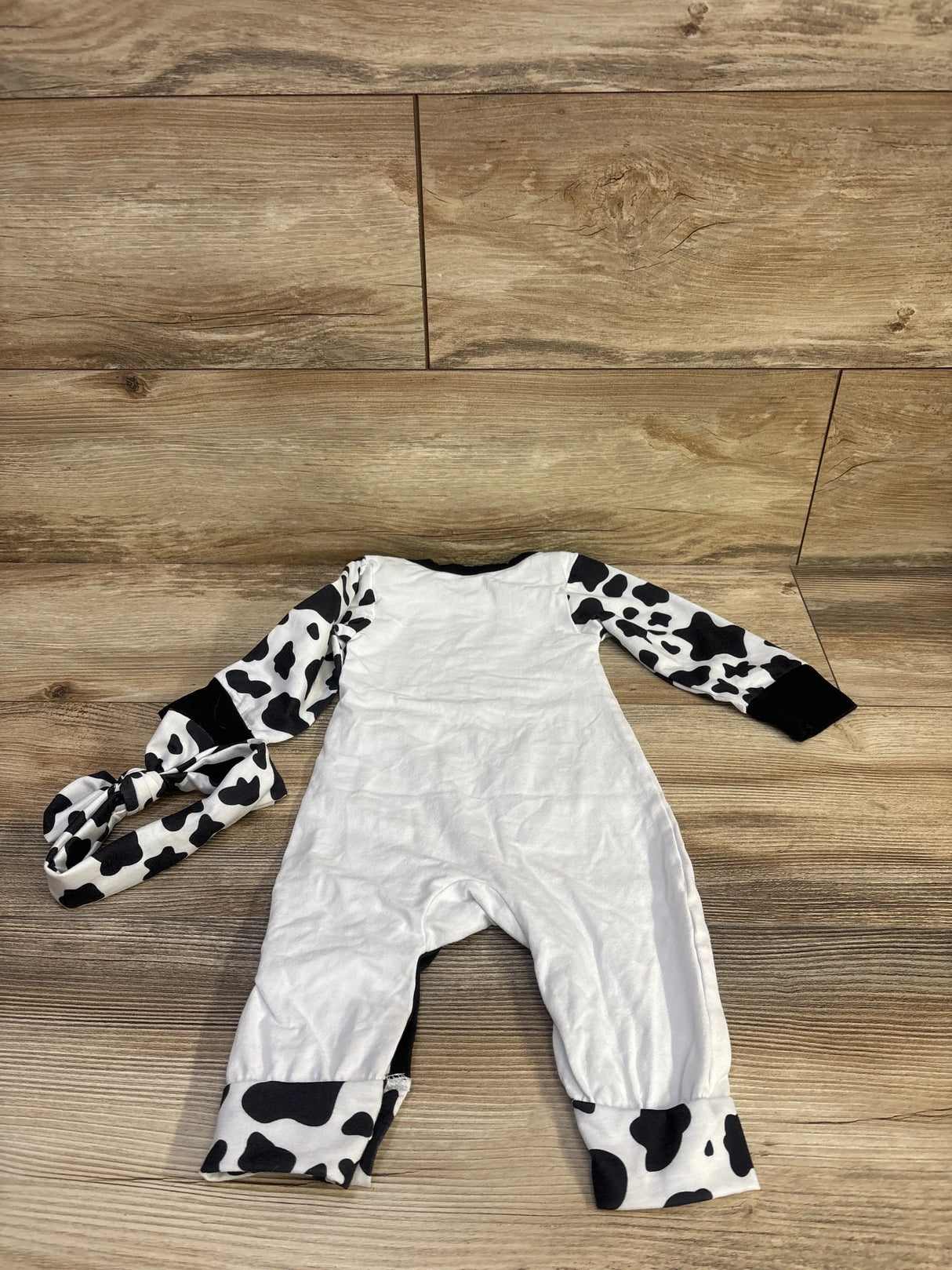 Top down view of white baby coverall and headband, 12-18 months, showing black cow print sleeves and cuffs on wood floor