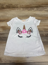 Front view of a white Btween unicorn shirt showcasing a floral unicorn face graphic and short sleeves on a wood background