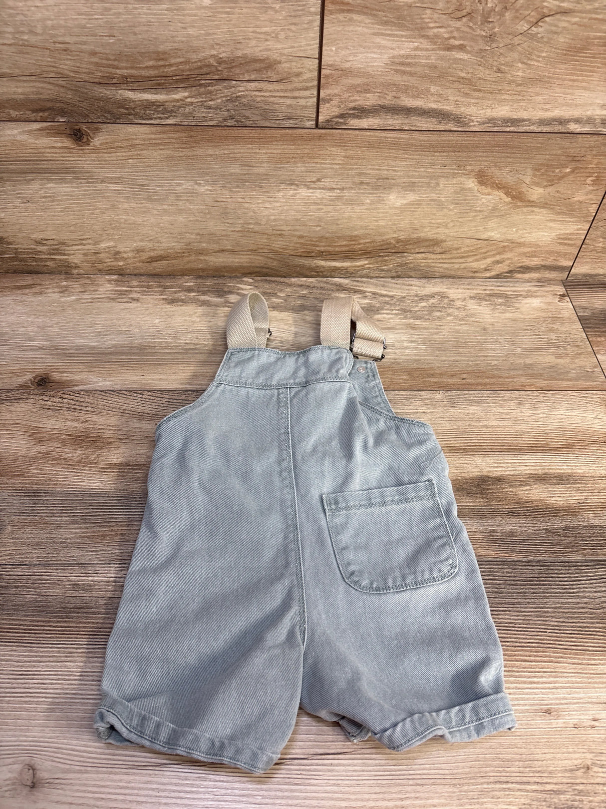 Flat lay front view of toddler denim shortalls showcasing adjustable tan straps, single front patch pocket and rolled hems on wood background