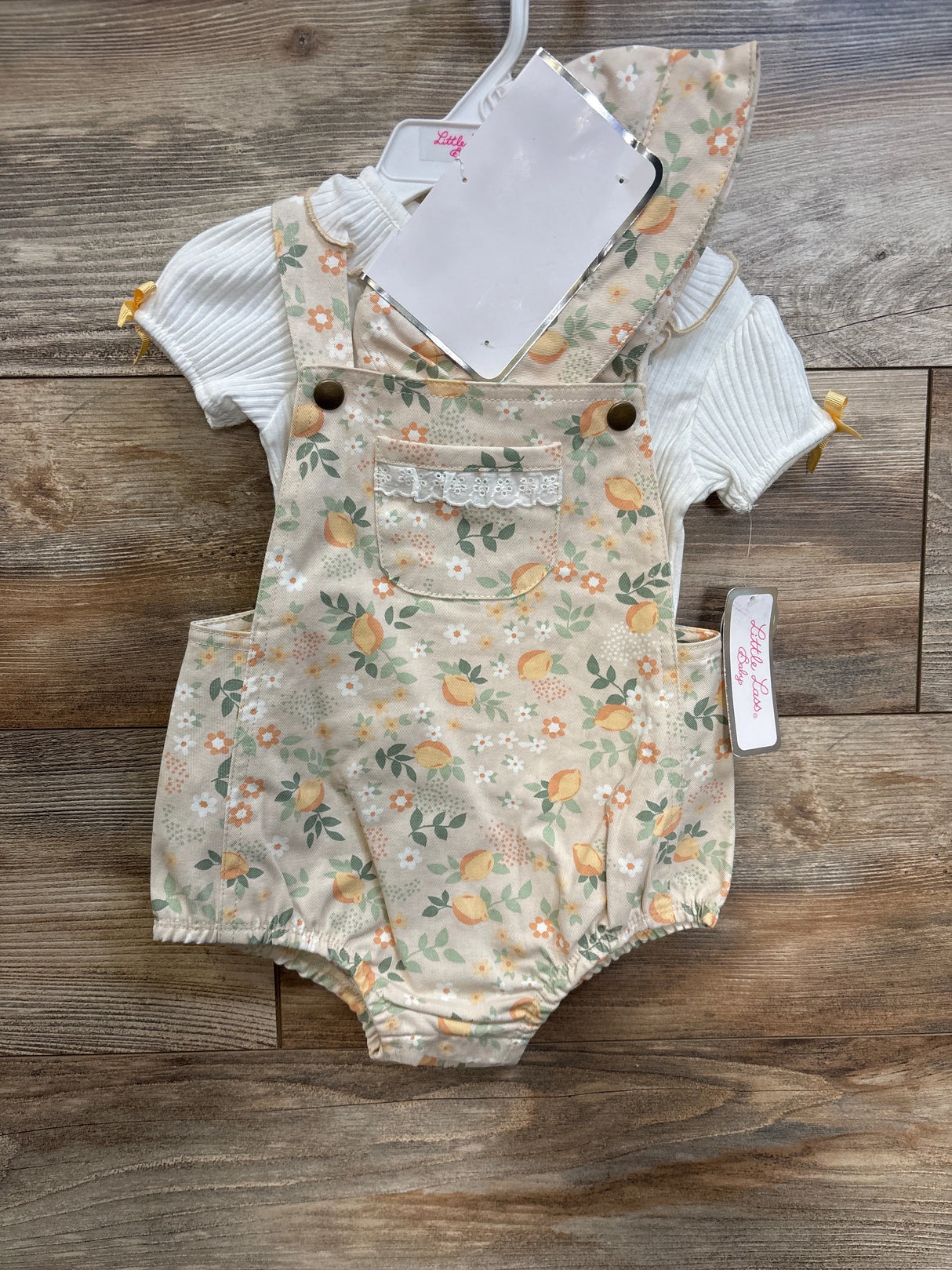 Flat lay of baby floral shortall romper and white ribbed shirt showing front pocket with lace trim, brass button straps, elastic leg openings on wood background