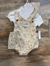 Flat lay of baby floral shortall romper and white ribbed shirt showing front pocket with lace trim, brass button straps, elastic leg openings on wood background