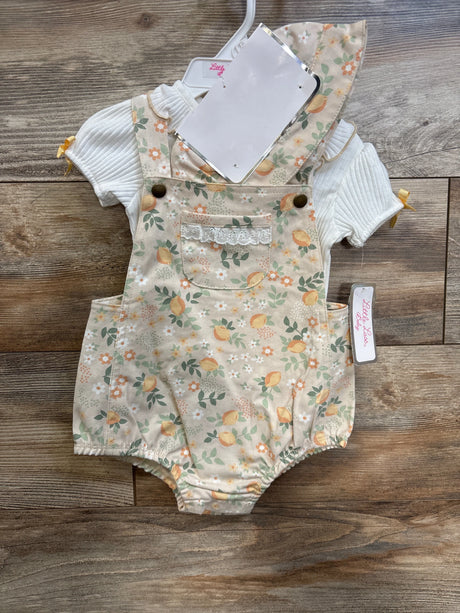Flat lay of baby floral shortall romper and white ribbed shirt showing front pocket with lace trim, brass button straps, elastic leg openings on wood background