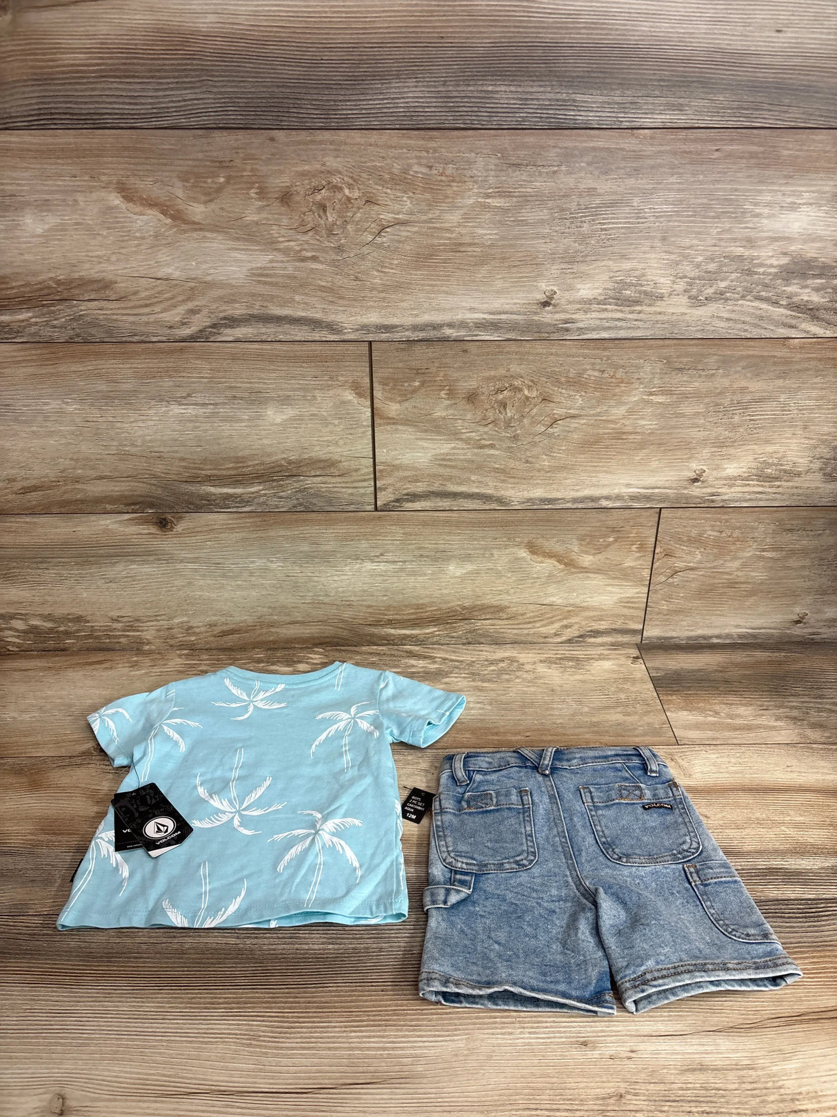 Top-down view of Volcom toddler two-piece set showcasing a light-blue palm-tree short-sleeve shirt and stonewashed jean shorts on wood floor