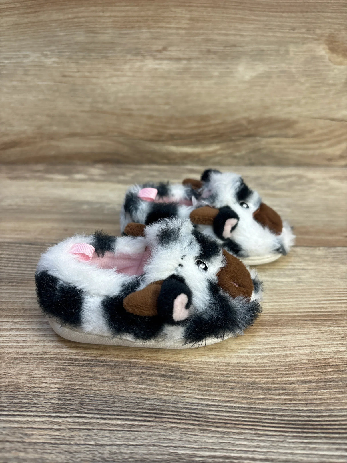 Plush cow print baby slippers with cute animal face and pink interior on wood background