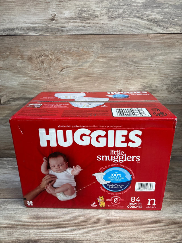 Front view of Huggies Little Snugglers 84-count newborn diaper box showing HUGGIES logo, baby, and protection callout on wood background