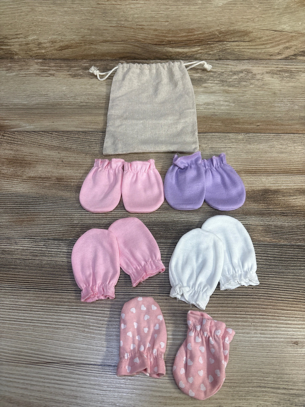 Top-down view of five pairs of baby mittens (pink, lavender, white, pink hearts) with beige pouch on wood background