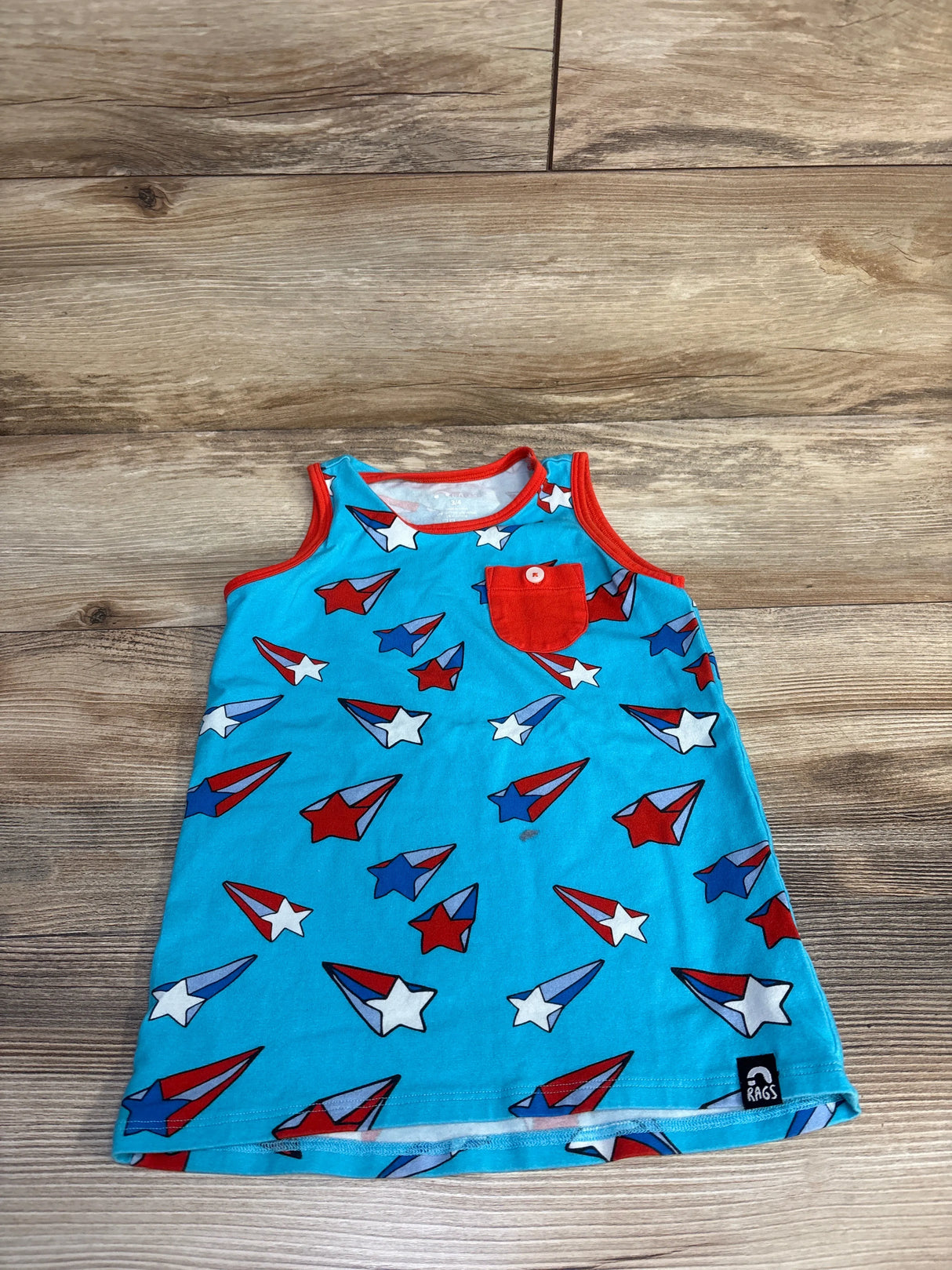 Flat lay of light-blue toddler tank top with red trim and pocket, red-white-blue star and airplane print on wood background