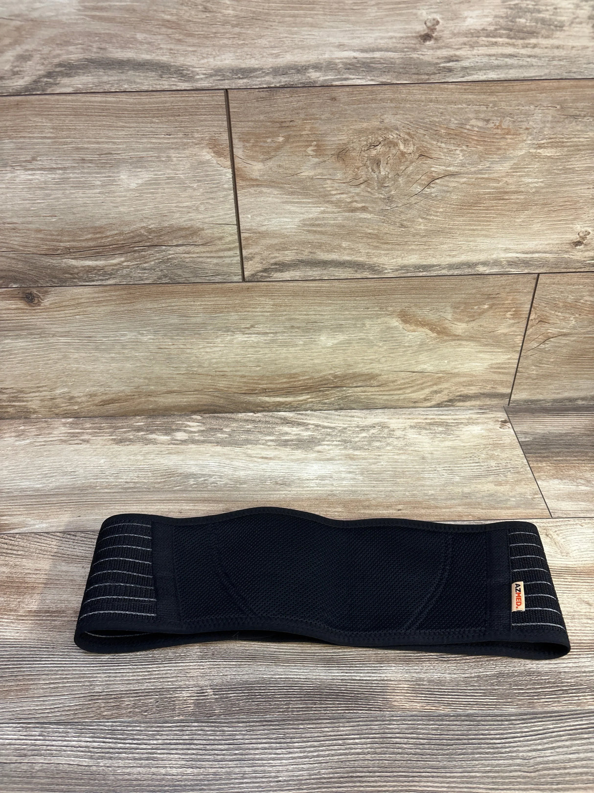 Flat-lay of AZMed maternity back support band in black showing breathable mesh center and adjustable Velcro straps on wood floor background