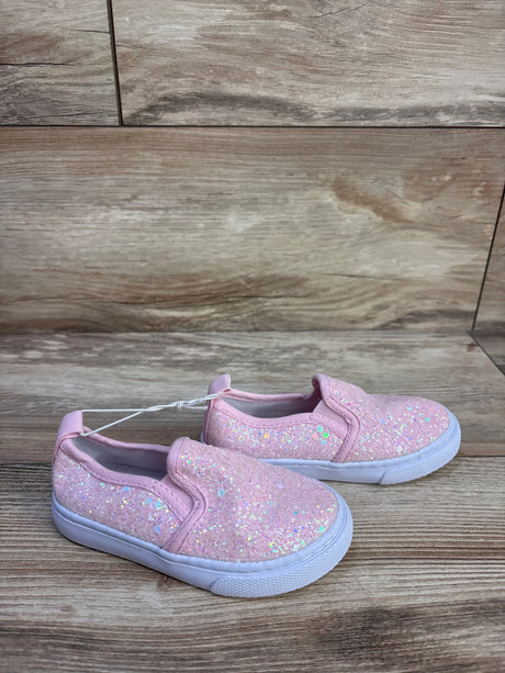 Angled side view of Cat and Jack pink slip-on toddler sneakers showing iridescent glitter uppers and white soles on wood background