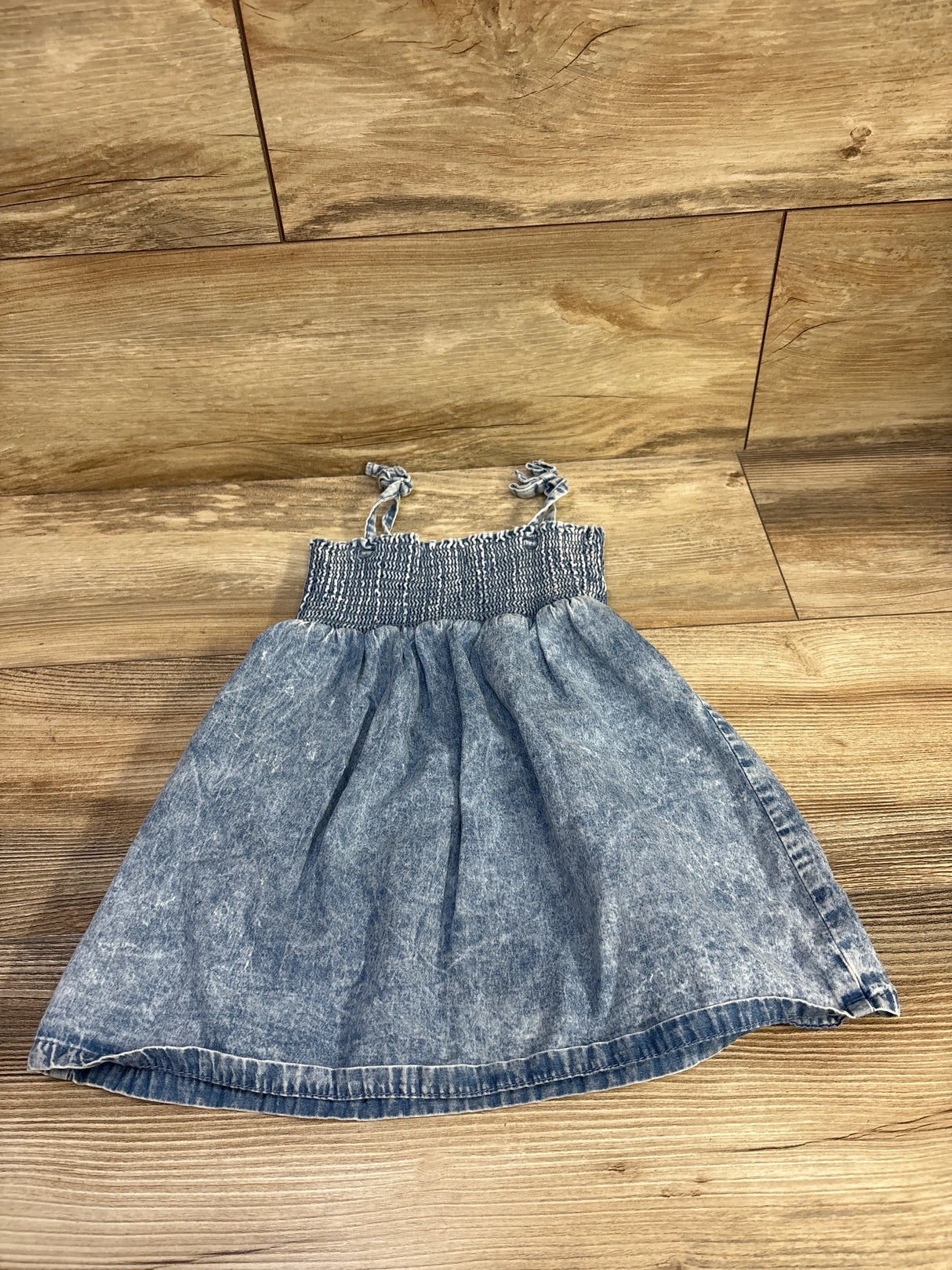 Flat lay of blue chambray toddler dress size 24 months showcasing smocked bodice and tie shoulder straps on wood floor