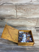 Top view of open Dowdle Best of the World 100-piece puzzle box with loose colorful pieces on wood background