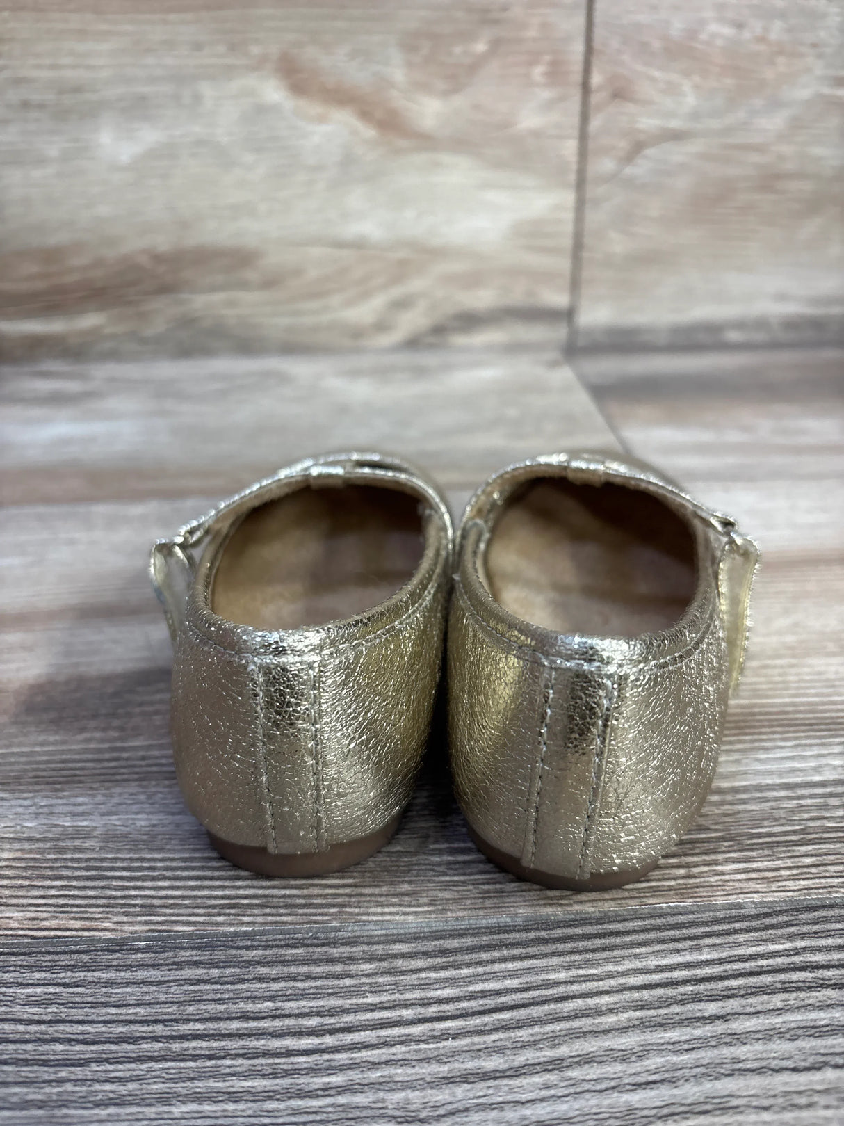 Rear view of Jumping Beans Susie T-Strap ballet flats showcasing metallic gold textured finish and stitched heel with wood-grain background