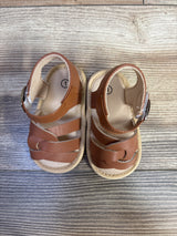 Top-down view of brown baby soft-sole sandals showing ankle strap with buckle and braided front straps on light wood floor