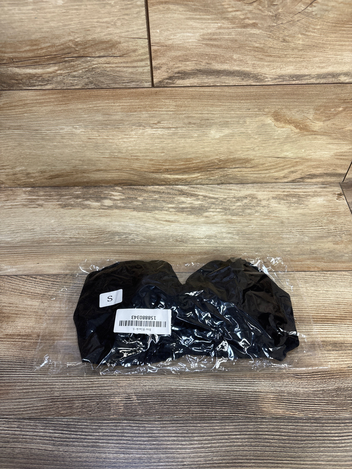 Top-down view of black nursing bra folded in clear plastic packaging showing size S sticker and barcode on wood floor background