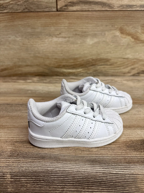 Side view of Adidas Superstar EL I sneakers showcasing white uppers, laces, and three stripes on a wood floor
