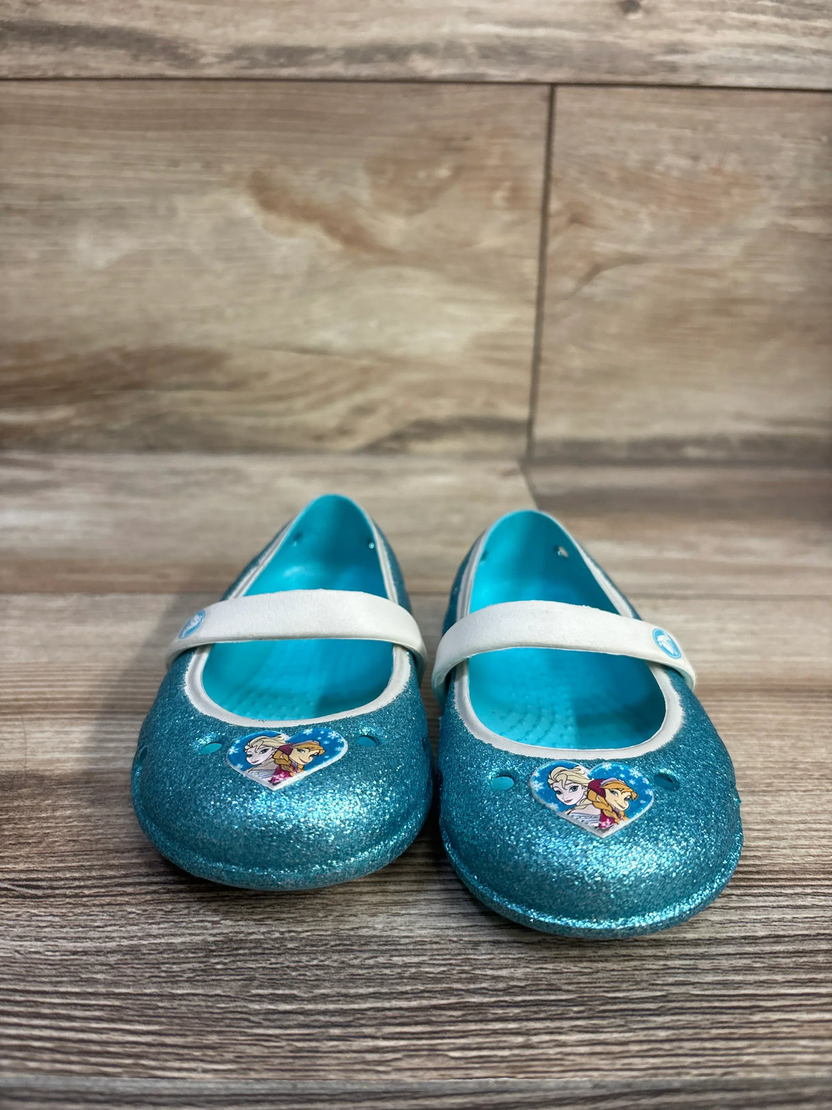 Front view of toddler blue glitter Crocs Mary Jane flats showcasing white strap and Frozen character heart stickers on a wood plank background