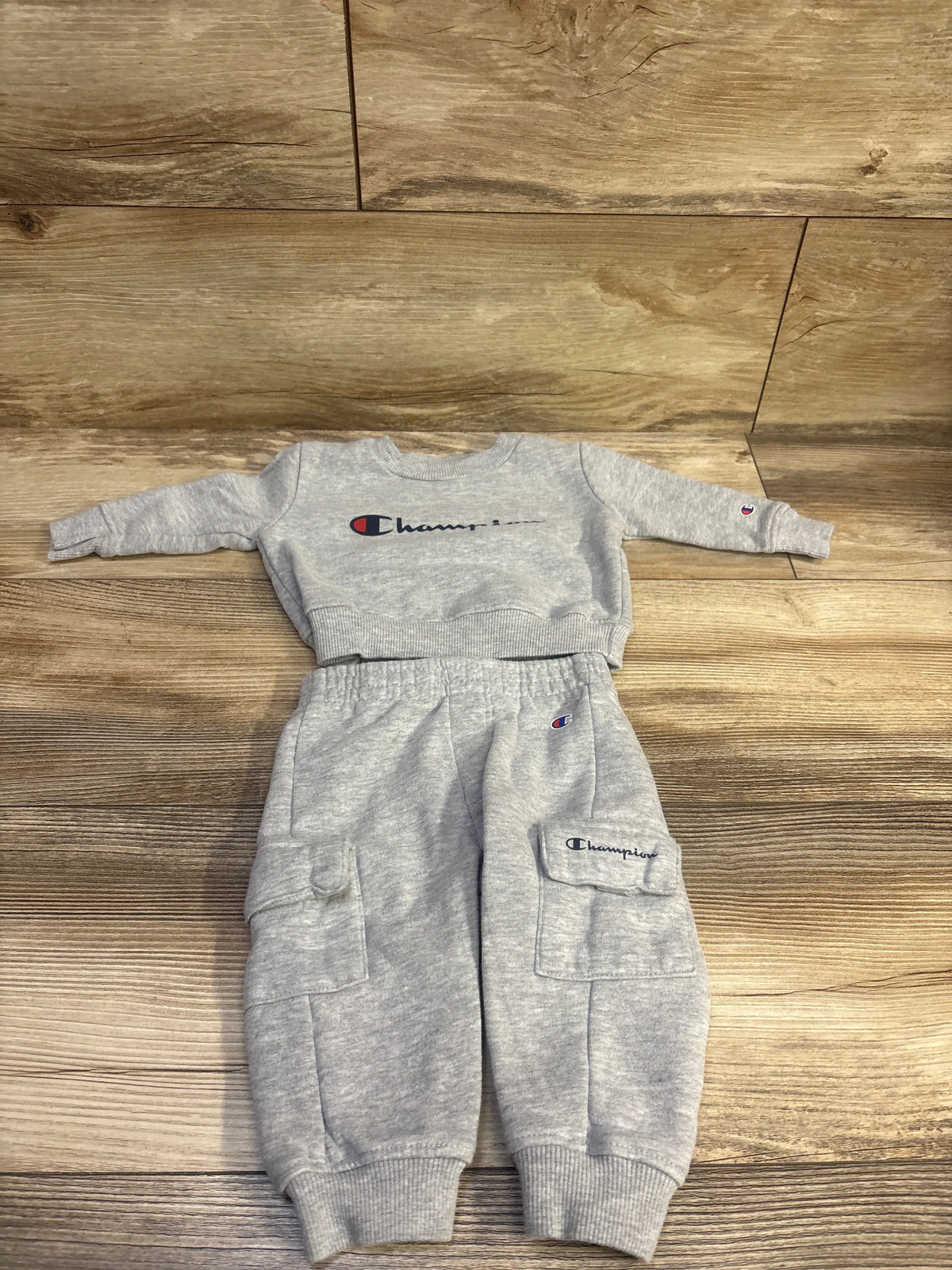 Flat lay of a grey Champion 2-piece pullover sweatshirt and joggers set in size 3 months with cargo pockets and ribbed cuffs on a wood floor