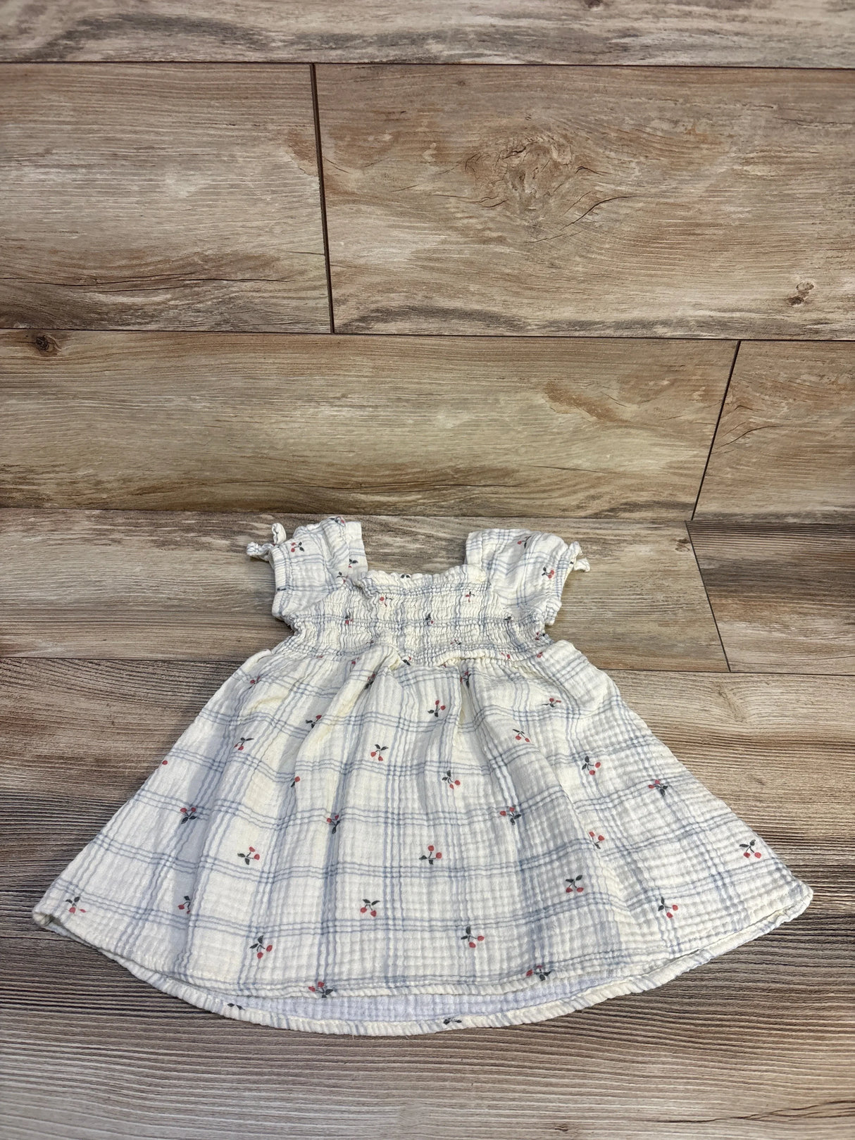 Flat lay of ivory smocked muslin puff-sleeve baby dress with smocked bodice, blue check and red floral print on wood