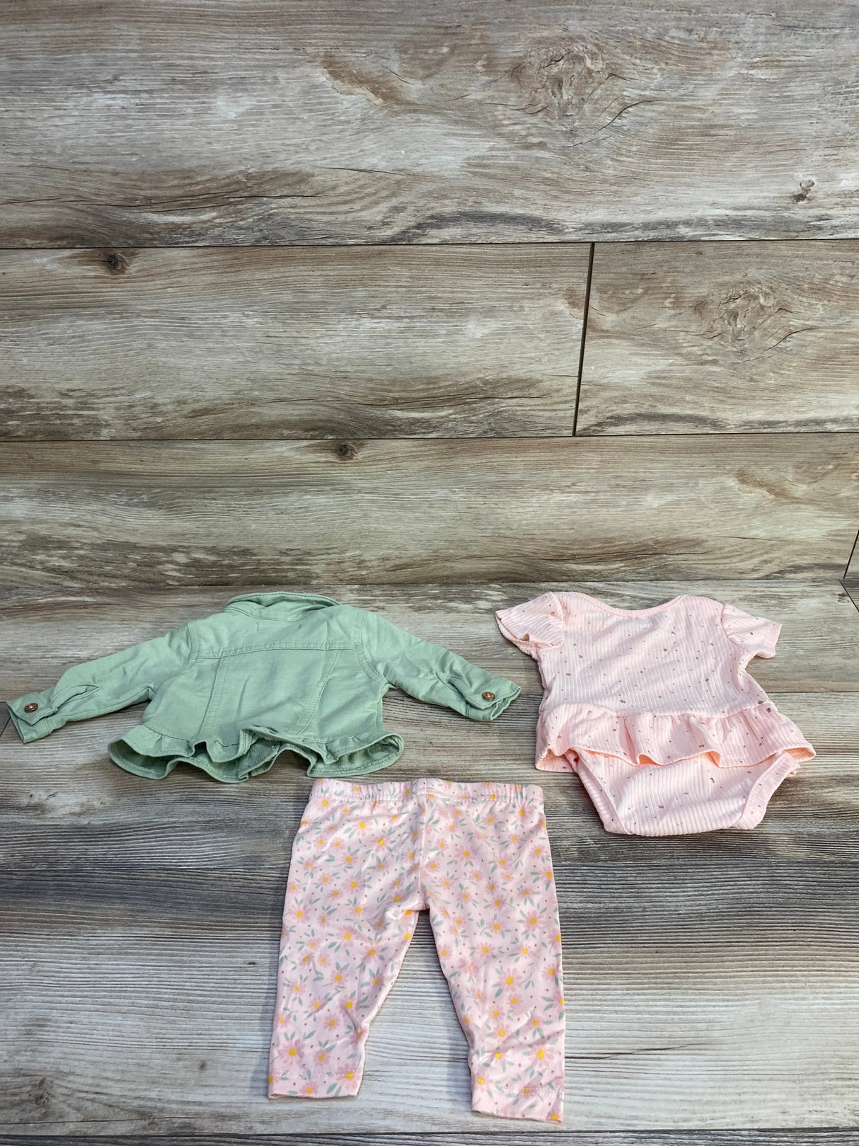Flat lay of Nanette Baby 3-piece newborn set (size 0-3M) showing green jacket, pink ruffled bodysuit, and pink floral pants on wood background