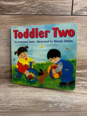 Angled front of Toddler Two board book showing illustrated cover with two children, a ball and small dog on grass against wood background