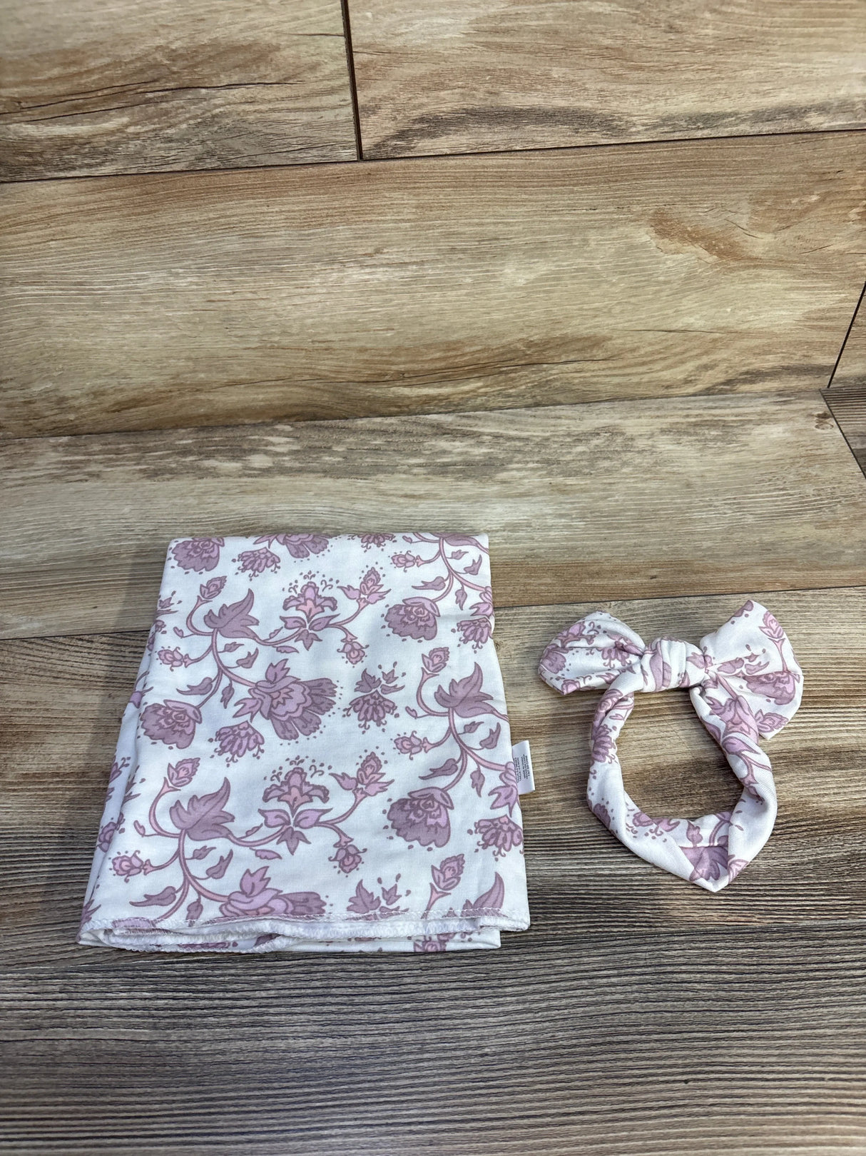 Top-down view of 2-piece Toby Floral baby headband and wrap set showcasing pink floral print on white fabric against wood-grain background