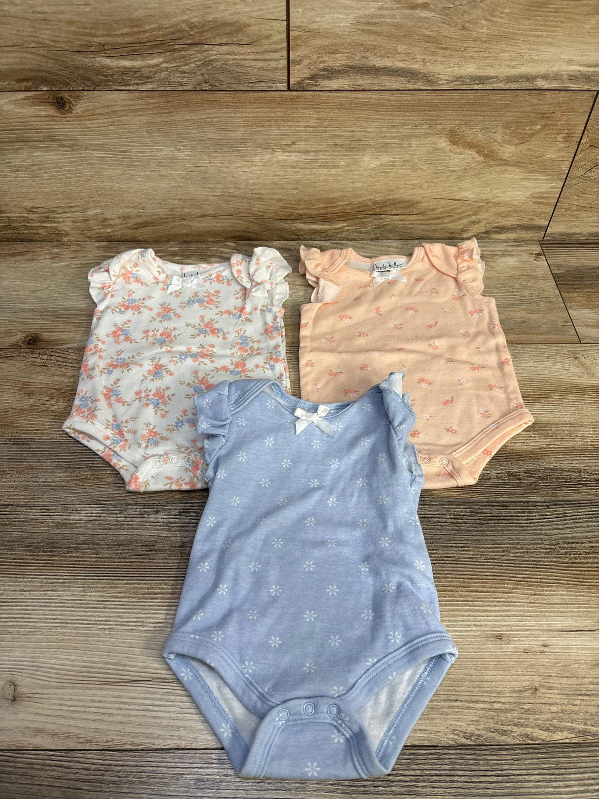 Top view of three flutter-sleeve baby bodysuits (white floral, peach, light blue) with ruffle sleeves and snap closures on wood background