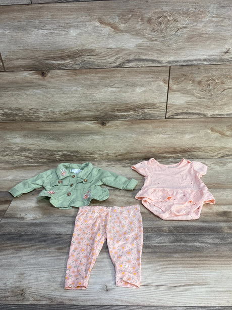 Flat-lay of Nanette Baby 3-piece set: embroidered green jacket, pink ruffle bodysuit, and floral leggings on wood background