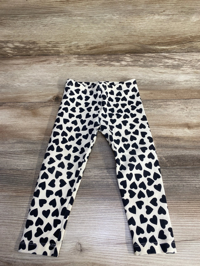 Flat lay of ivory girls' leggings showing all-over black heart print and elastic waistband on light wood floor, size 2T
